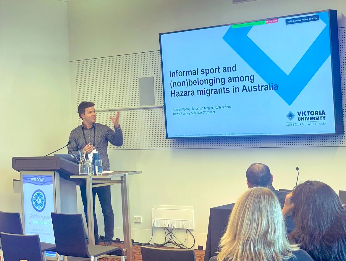 Ramon Spaaij Presents on Informal Sport and Nonbelonging among Hazara migrants in Australia standing at a lecture motioning with his hand toward the projected slides on the screen