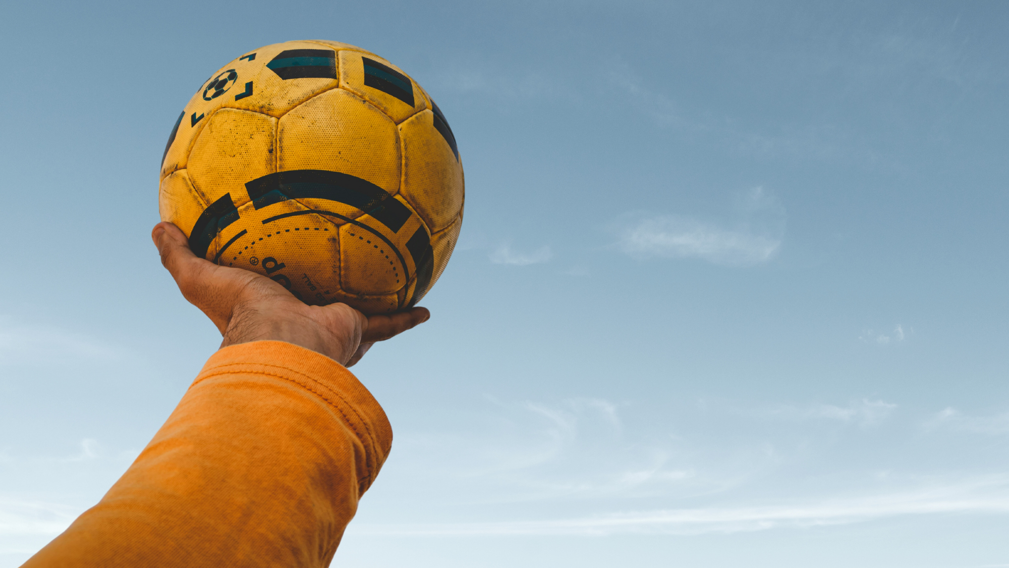 A person wearing a yellow shirt holds a soccer ball high to the sky. The soccer ball is yellow and black. The person is wearing a yel