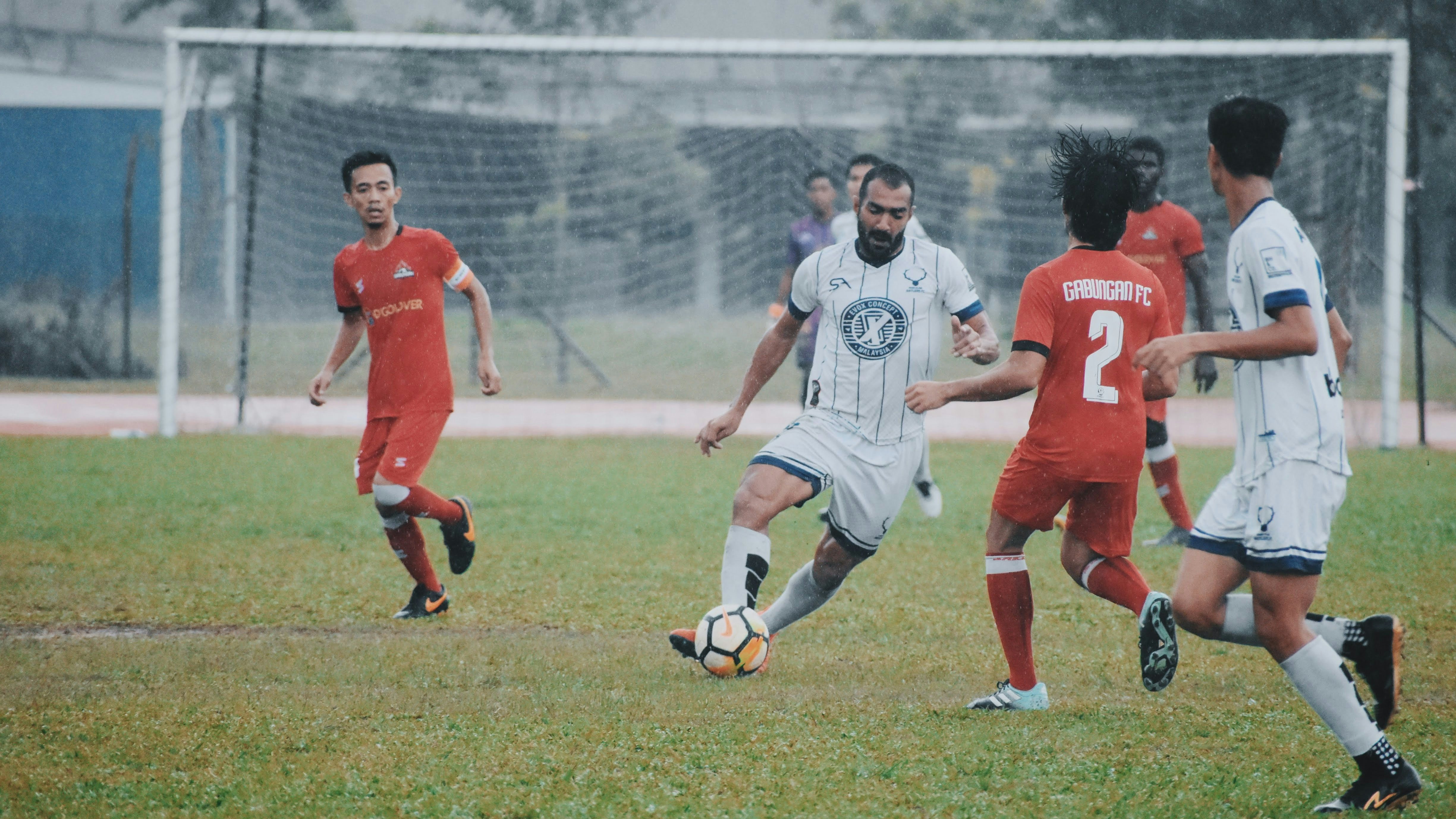Seven men on a soccer field. Three are on a team with red shirts. Three are on a team with white shirts. One man with a white shirt has control of the ball. A goalie in a purple shirt is in the distance protecting the goal.