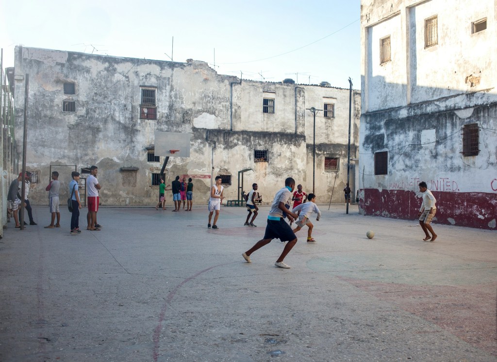 Several children in Havanna, Cuba kick a soccer ball in a courtyard. People relax and look on from the sidelines. There are concrete buildings on either side.