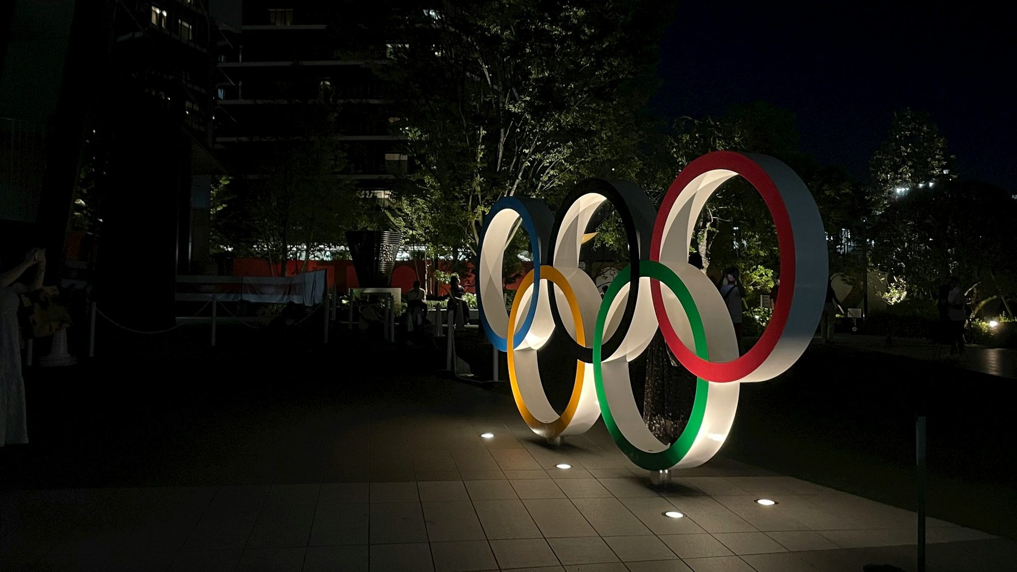The olympic rings in blue, yellow, black, green, and red at night underlit by 5 in-ground lights in a clearing in Tokyo for the 2020 Olympics